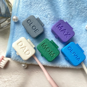 Può includere: Una collezione di copri spazzolini colorati, ognuno con un nome o titolo in rilievo. I coperchi sono nei toni del grigio, viola, verde, blu e bianco. I nomi includono "CLOE", "MAX", "DAD", "MOM" e "OLIVIA".