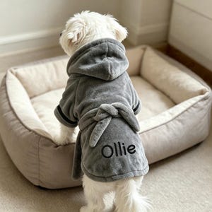 May include: A small white dog wearing a plush grey hooded bathrobe with a tie waist. The robe features black trim on the sleeves and the name Ollie embroidered in black thread on the back. The garment is made of a soft, fuzzy material.