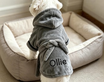 May include: A small white dog wearing a plush grey hooded bathrobe with a tie waist. The robe features black trim on the sleeves and the name Ollie embroidered in black thread on the back. The garment is made of a soft, fuzzy material.