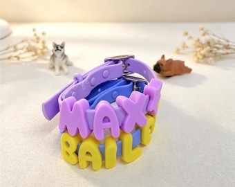 May include: A silicone bracelet featuring two stacked bands in light purple and blue. The top band displays the purple raised letters MAXI, while the bottom band features yellow raised letters spelling BAILEY. The bracelet includes a metal buckle.