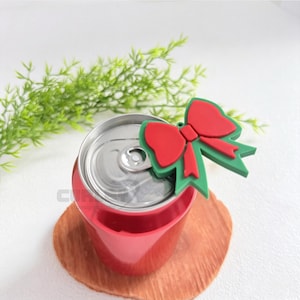 May include: A red beverage can sleeve featuring a decorative silicone topper shaped like a red bow with green trim. The topper is positioned over the silver pull tab of the aluminum can, which is encased in the solid red cylindrical holder.