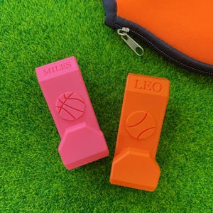 May include: Two personalized basketball-themed phone stands. One is pink with "MILES" and a basketball design, the other is orange with "LEO" and a tennis ball design. The stands are on a green surface.