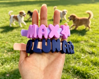 May include: Two silicone beaded wristlets featuring raised block letters. The top lavender band displays the name MAX with a star icon, while the bottom peach-colored band displays the name LUNA with a flower icon, both made of dark blue letters.