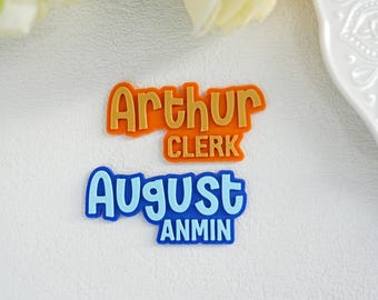 May include: Two name tags with text. The top tag is orange with the name "Arthur" and the word "Clerk" below it. The bottom tag is blue with the name "August" and the word "Anmin" below it. The tags are on a white surface.