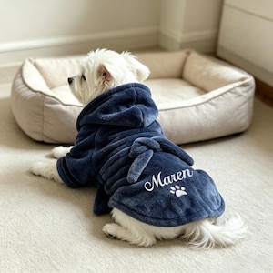 May include: A small white dog wearing a navy blue plush hooded bathrobe with a tie belt. The back of the robe features the embroidered white name Maren above a small white paw print graphic. The dog is positioned in front of a beige pet bed.