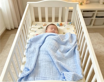 May include: A baby sleeping in a white wooden crib, covered by a light blue muslin blanket with a textured, crinkled finish. The baby lies on a cream-colored mattress featuring a subtle pattern of small, colorful animal illustrations and shapes.