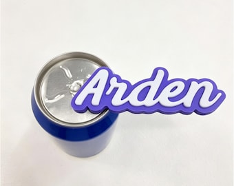 May include: A blue aluminum beverage can with a purple and white silicone tab topper featuring the name Arden in a cursive script font. The topper is attached to the silver pull tab of the can, extending horizontally to the right side.