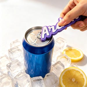 May include: A purple silicone can opener tool with the white text "Archie" embossed on the handle, positioned to lift the tab of a blue aluminum beverage can surrounded by clear ice cubes and two bright yellow lemon slices.