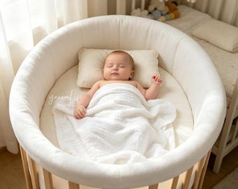May include: A baby sleeping in a round wooden crib with a white mattress, a small rectangular pillow, and a white muslin blanket. The crib features vertical wooden slats and a soft, padded white bumper. The text yanilife Studio is visible.