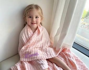 May include: A soft, light pink muslin blanket with a textured, crinkled fabric finish. The blanket features the name Lucas embroidered in a purple cursive script on the front. The lightweight material is draped loosely over the shoulders.