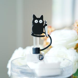 May include: A black cat-shaped bottle stopper with a white knife, a black handle, and a small charm with the letter "P". The stopper is on a clear container with white flowers in the background.
