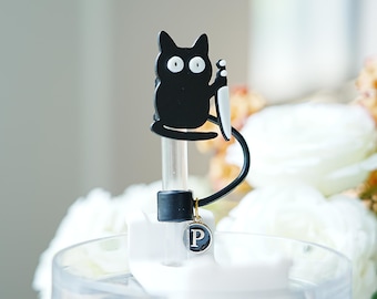 May include: A black cat-shaped bottle stopper with a white knife, a black handle, and a small charm with the letter "P". The stopper is on a clear container with white flowers in the background.