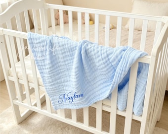 May include: A light blue muslin baby blanket with a crinkled texture draped over a white wooden crib. The blanket features the name Naylani embroidered in a dark blue cursive script on the lower corner of the fabric, adding a personalized touch.