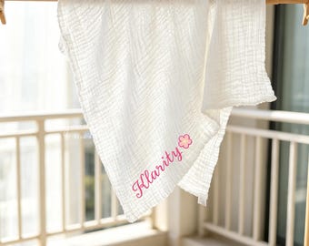 May include: A white muslin swaddle blanket featuring a crinkled texture and a soft, lightweight appearance. The fabric is draped over a wooden rod and displays the name Klarity in pink cursive script, accompanied by a small pink flower icon.