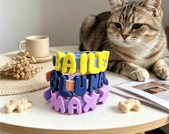 May include: A stack of three silicone pet collars in light blue, peach, and purple. The collars feature large, raised, colorful letters spelling out the names BAILEY, LUNA, and MAX, with a paw print icon included on the middle collar.