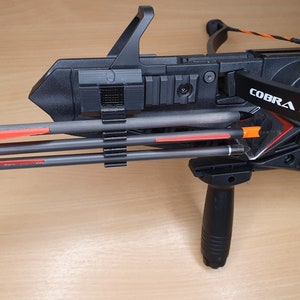 May include: A black crossbow with a scope and a black and orange string. The crossbow has a black pistol grip and a black arrow holder with three arrows. The arrows have orange fletching.