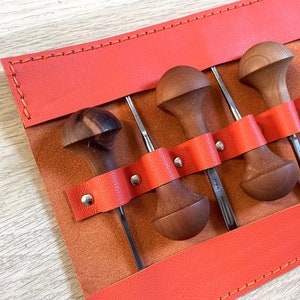 May include: A set of four wood carving tools with stainless steel blades, each with a unique wooden handle. The tools are held in place by a bright orange leather strap within a tan leather tool roll. The roll is stitched with orange thread.