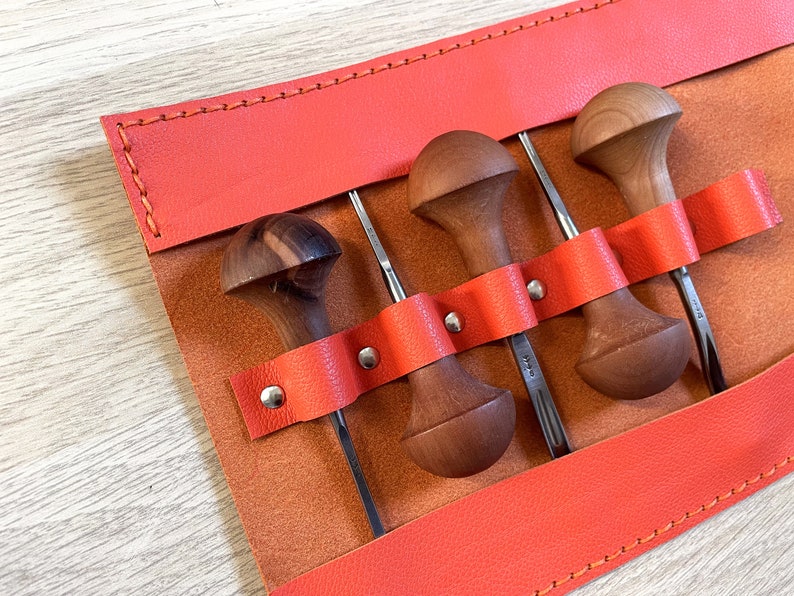 May include: A set of four wood carving tools with stainless steel blades, each with a unique wooden handle. The tools are held in place by a bright orange leather strap within a tan leather tool roll. The roll is stitched with orange thread.