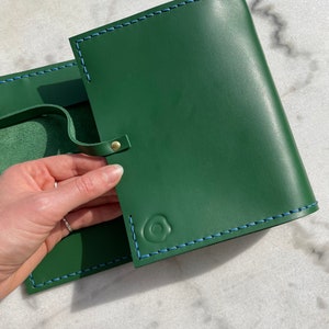May include: Green leather journal with blue stitching and a snap closure. The journal has a loop for a pen and a debossed design on the front. The journal is open, revealing the interior pages.