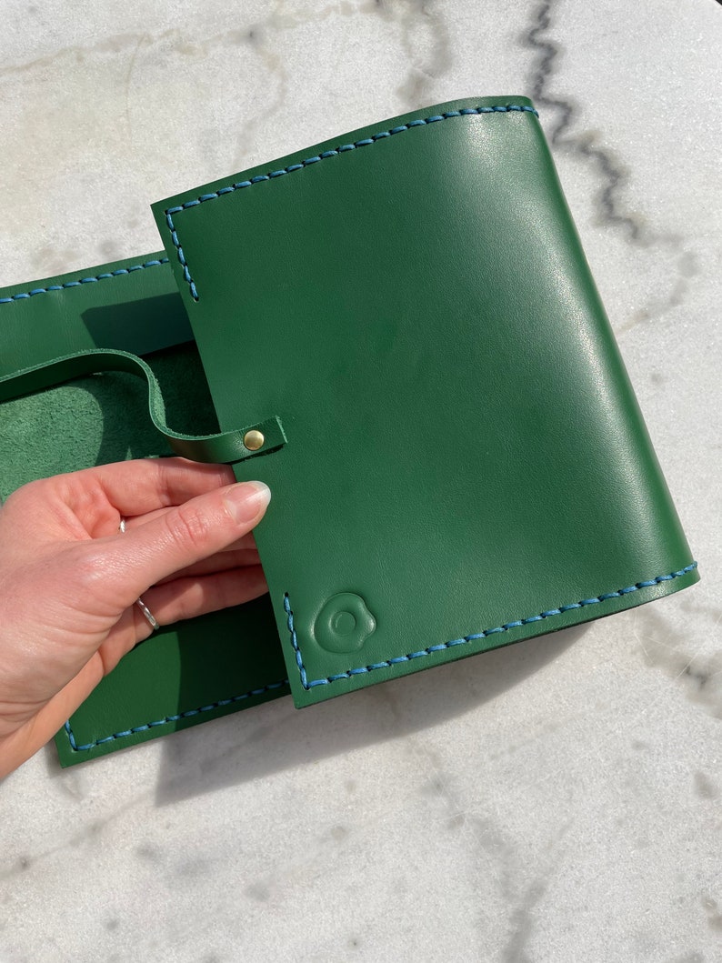 May include: Green leather journal with blue stitching and a snap closure. The journal has a loop for a pen and a debossed design on the front. The journal is open, revealing the interior pages.