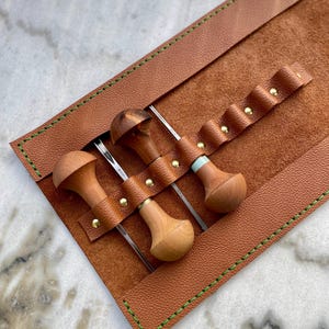 May include: A leather tool roll in a warm brown hue, with green stitching and brass rivets. It holds three wood-handled tools with metal blades. The tools have unique wooden handles. The roll is open, displaying the tools.