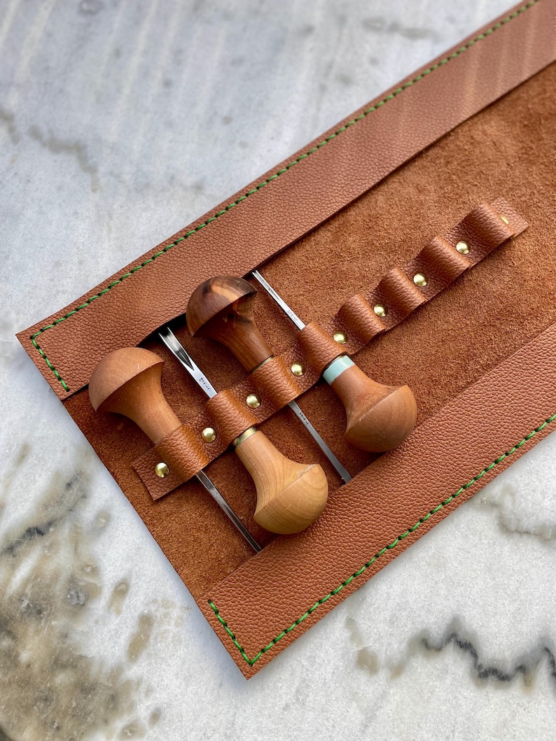 May include: A leather tool roll in a warm brown hue, with green stitching and brass rivets. It holds three wood-handled tools with metal blades. The tools have unique wooden handles. The roll is open, displaying the tools.