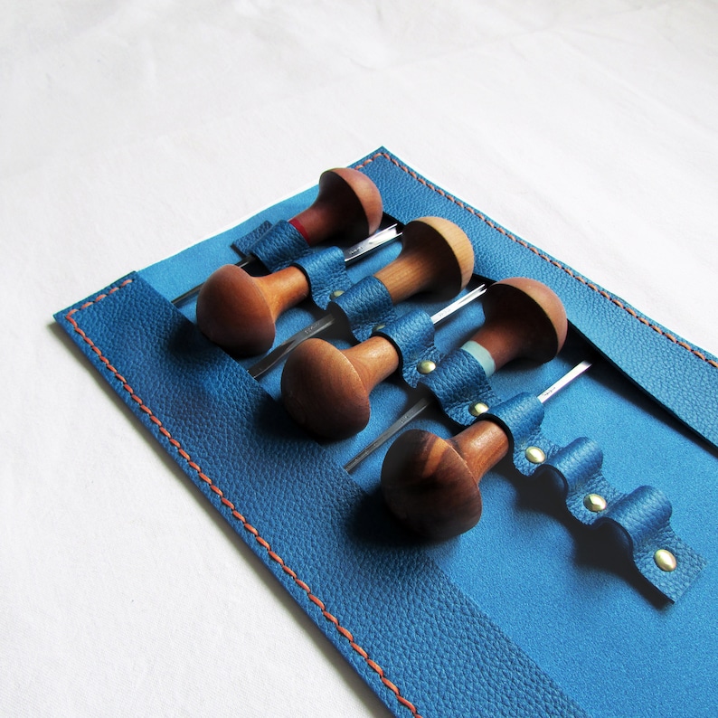 May include: A set of wood carving tools with wooden handles and metal blades, secured in a blue leather tool roll. The roll has orange stitching and brass rivets. The tools are for woodworking.