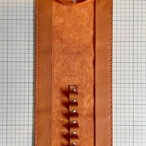 May include: An orange leather pen holder with a loop at the top and a series of small loops with rivets down the center. The holder is rectangular with stitched edges and a textured interior.
