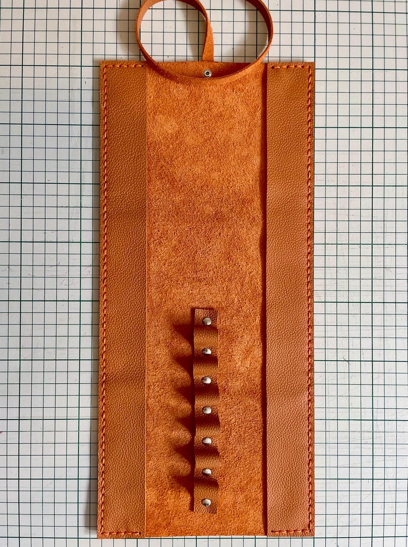 May include: An orange leather pen holder with a loop at the top and a series of small loops with rivets down the center. The holder is rectangular with stitched edges and a textured interior.