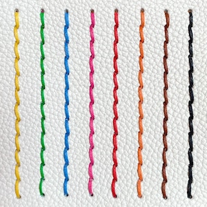 May include: Close-up of a white leather surface with nine vertical rows of colorful stitching. The stitches are in yellow, green, blue, pink, red, orange, brown, and black. The stitches are evenly spaced and create a textured pattern.