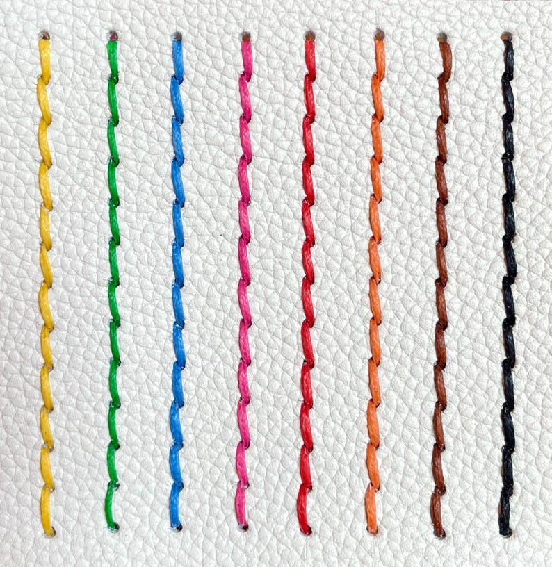 May include: Close-up of a white leather surface with nine vertical rows of colorful stitching. The stitches are in yellow, green, blue, pink, red, orange, brown, and black. The stitches are evenly spaced and create a textured pattern.