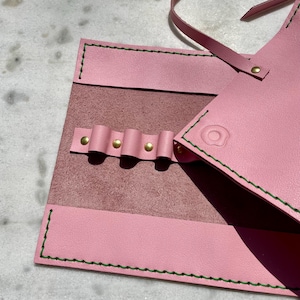 May include: A pink leather tool roll with green stitching and brass rivets. The roll is open, revealing a darker pink interior with loops for holding tools. A strap extends from the top for securing the roll.