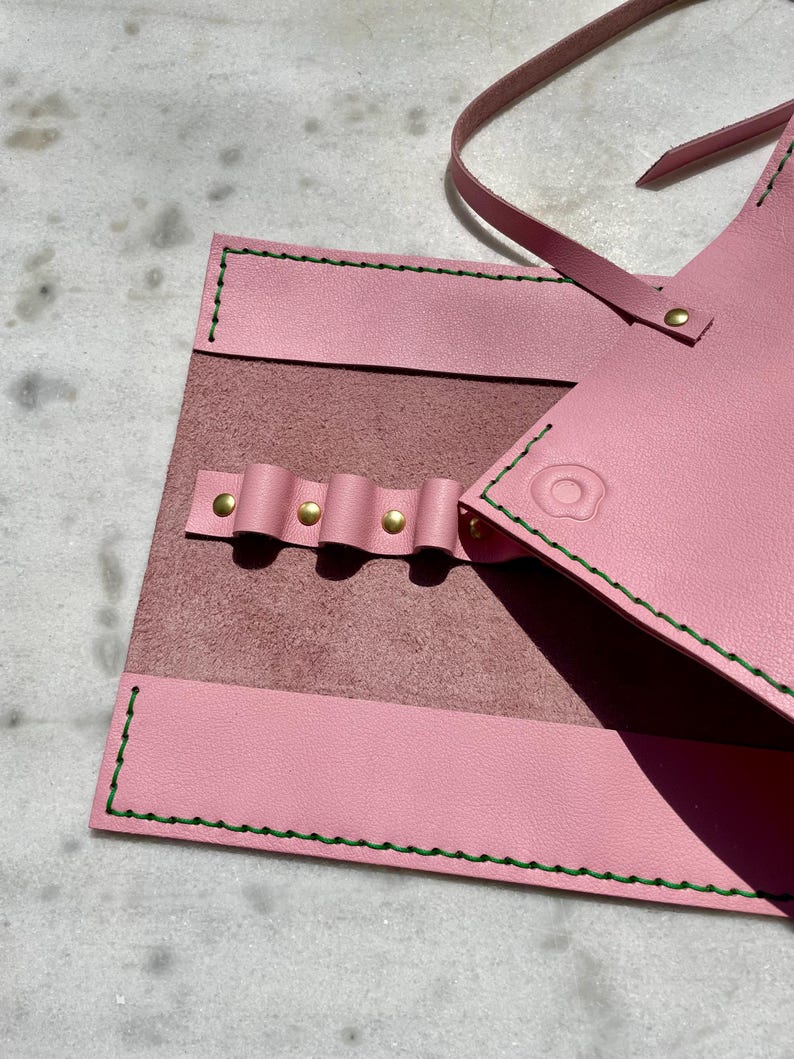 May include: A pink leather tool roll with green stitching and brass rivets. The roll is open, revealing a darker pink interior with loops for holding tools. A strap extends from the top for securing the roll.