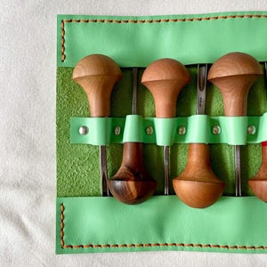 May include: A set of wood carving tools with wooden handles and metal blades, stored in a light green leather tool roll. The tool roll is stitched with brown thread.