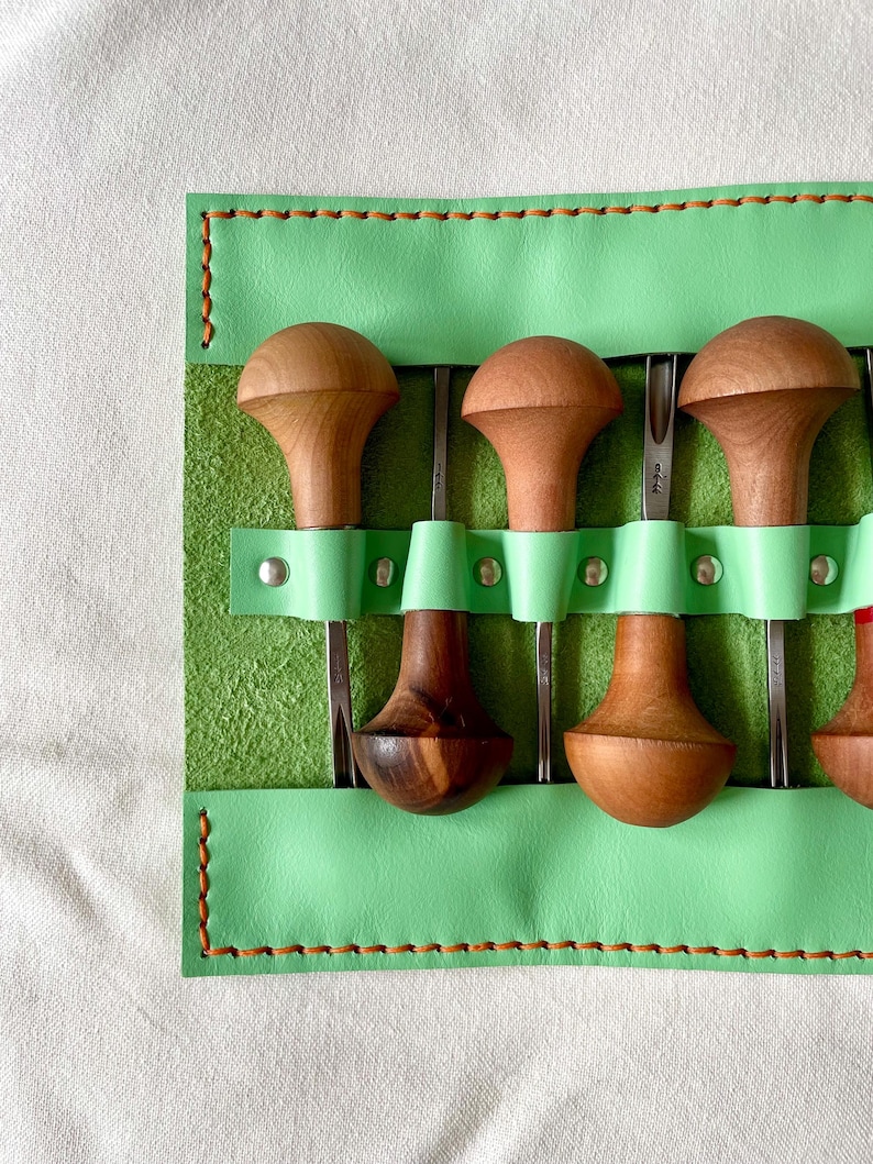 May include: A set of wood carving tools with wooden handles and metal blades, stored in a light green leather tool roll. The tool roll is stitched with brown thread.