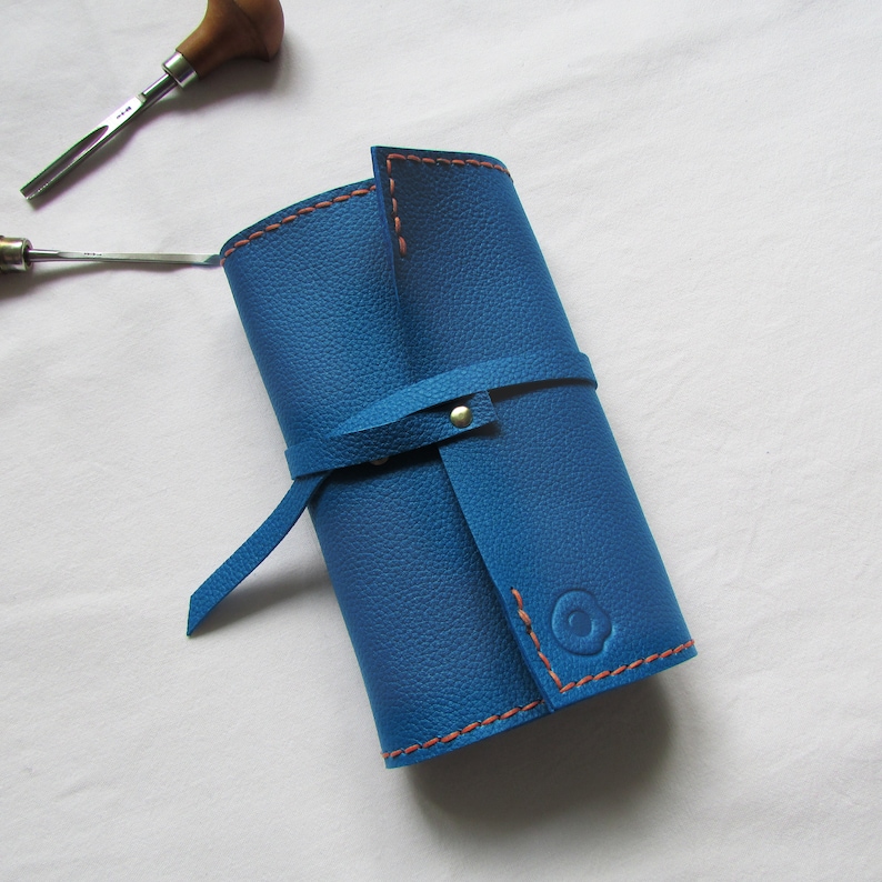 May include: A rolled-up blue leather tool roll with orange stitching and a strap closure. The roll is made of textured leather and has a circular embossed design on the front. A wood-handled carving tool is visible in the background.