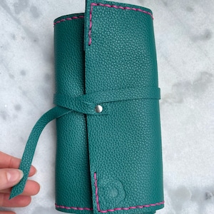 May include: Teal leather roll-up case with pink stitching. The case is closed with a matching teal strap and a silver snap. The leather has a textured appearance. The case is designed to hold art supplies.