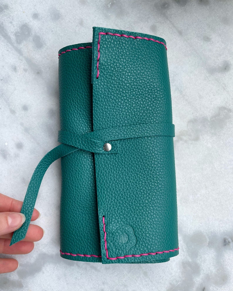 May include: Teal leather roll-up case with pink stitching. The case is closed with a matching teal strap and a silver snap. The leather has a textured appearance. The case is designed to hold art supplies.