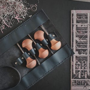 May include: A black leather tool roll holds four wood carving tools with wooden handles and metal blades. A metal ruler with centimeter markings lies above the roll. Pink shavings and a black and white architectural drawing are also present.