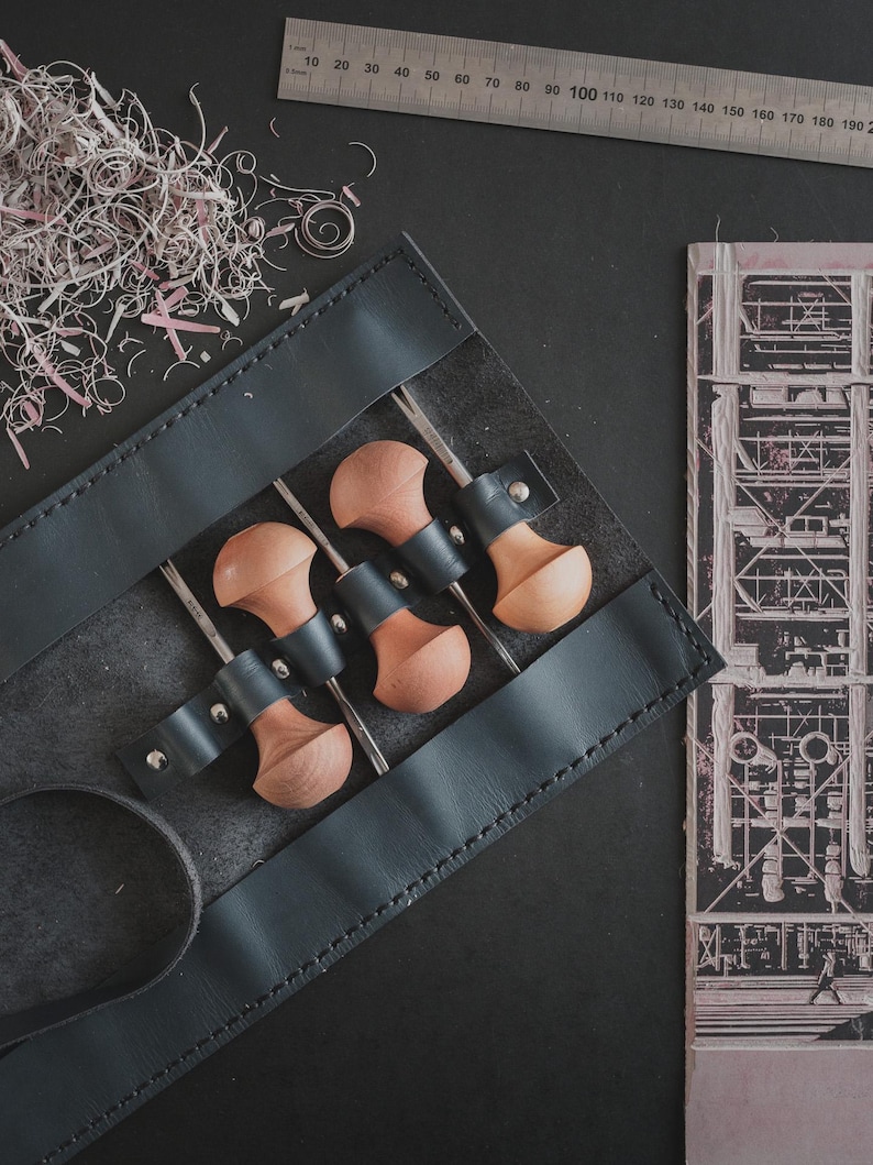 May include: A black leather tool roll holds four wood carving tools with wooden handles and metal blades. A metal ruler with centimeter markings lies above the roll. Pink shavings and a black and white architectural drawing are also present.