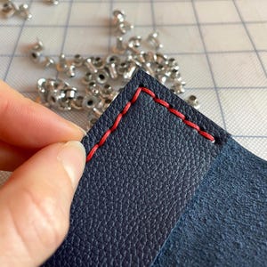 May include: Close-up of navy blue leather with red stitching. The leather is textured and the stitching is visible along the edges. Small silver rivets are scattered in the background, suggesting a crafting project.