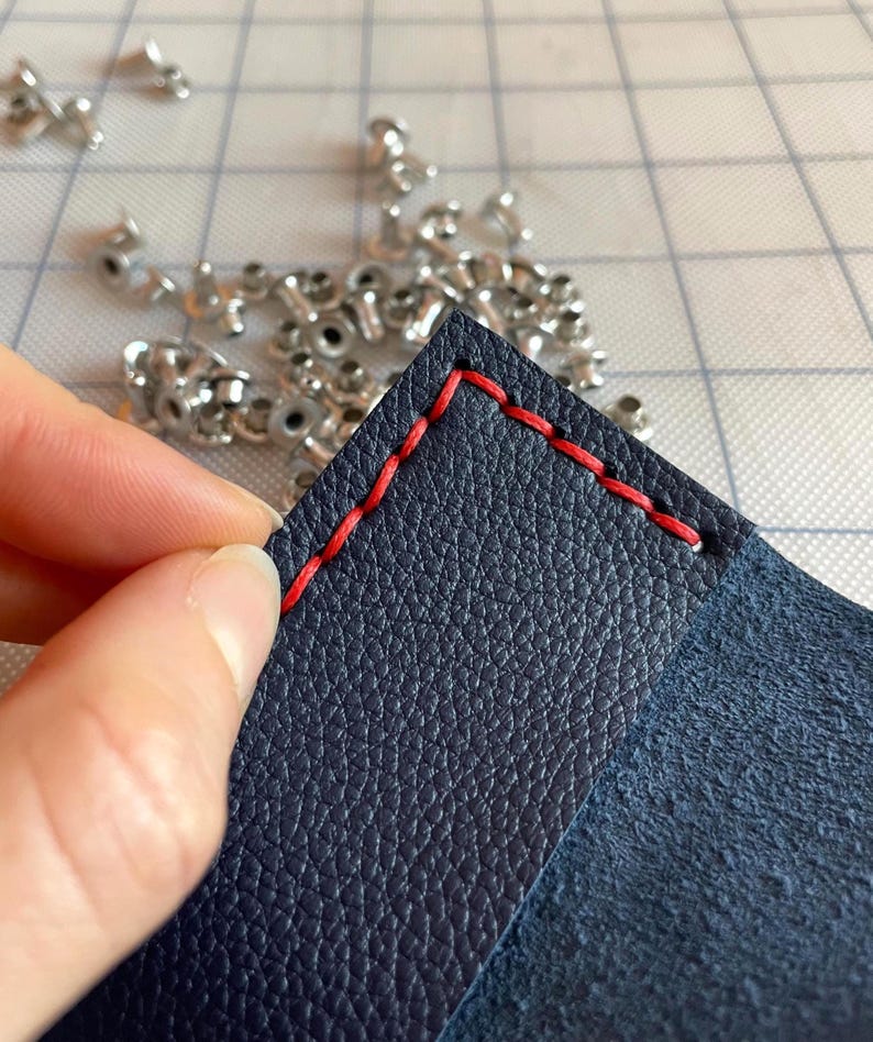 May include: Close-up of navy blue leather with red stitching. The leather is textured and the stitching is visible along the edges. Small silver rivets are scattered in the background, suggesting a crafting project.