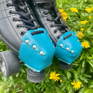 May include: Close-up of black Riedell roller skates with teal toe caps. The toe caps are studded with silver rivets. The skates are laced up with black laces and sit in a bed of green grass and yellow flowers.
