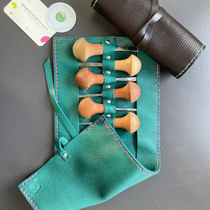 May include: A set of wood carving tools with wooden handles and metal blades, stored in a teal leather roll-up case with pink stitching. A rolled brown leather case with green stitching is also present. A business card and sticker are also visible.