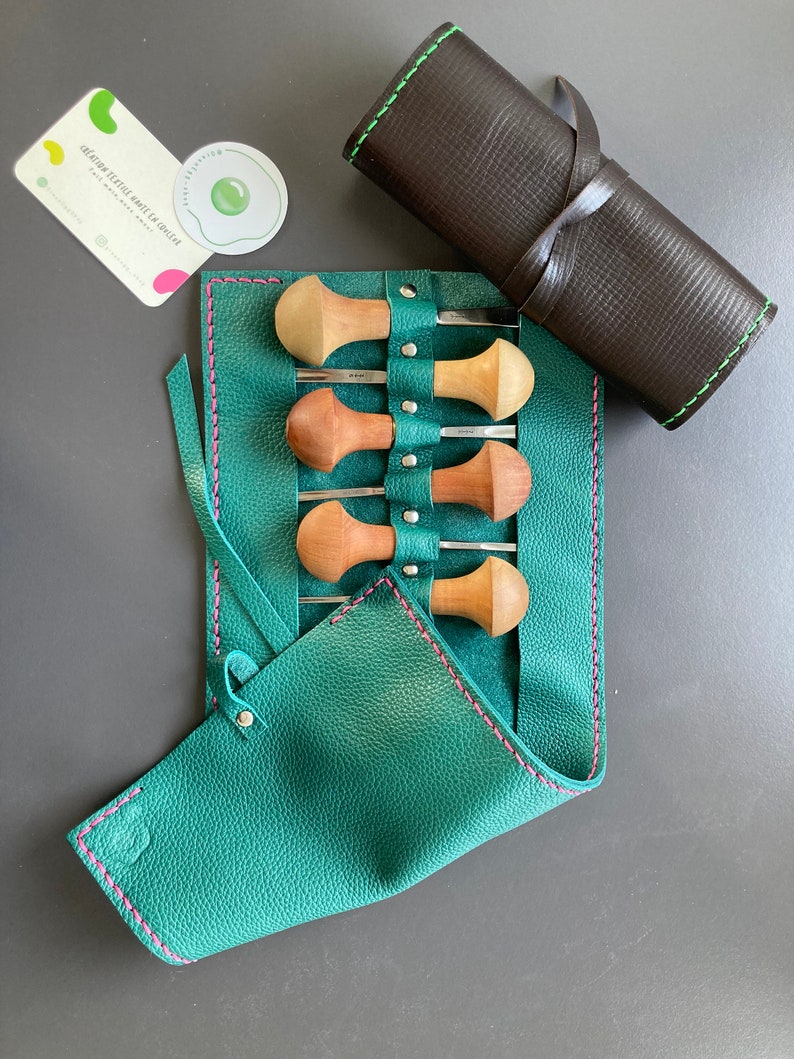 May include: A set of wood carving tools with wooden handles and metal blades, stored in a teal leather roll-up case with pink stitching. A rolled brown leather case with green stitching is also present. A business card and sticker are also visible.
