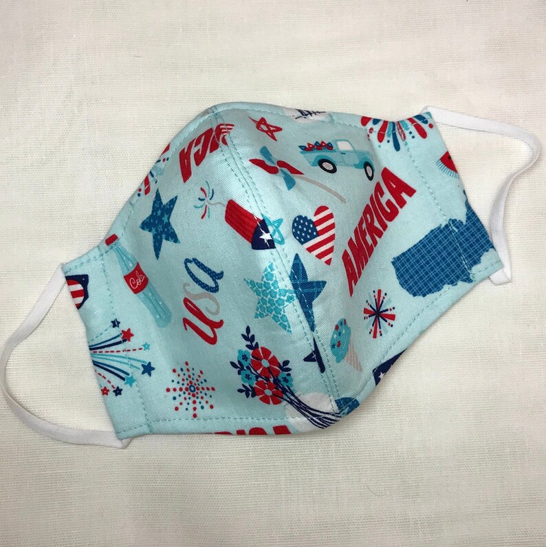 May include: A blue fabric face mask with a white elastic band. The mask has a pattern of red, white, and blue stars, fireworks, hearts, and the words "USA" and "America".