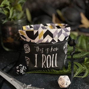 May include: Black and white dice bag with a geometric pattern and the text "This is how I roll". The bag is sitting on a table with a green plant and a dice in the foreground.