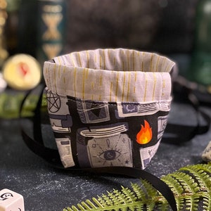 Puede incluir: Una bolsa de dados de tela negra y gris con forro blanco. La bolsa está decorada con un patrón de libros, ojos y llamas. La bolsa está sobre una superficie gris oscuro con un helecho verde y dados.