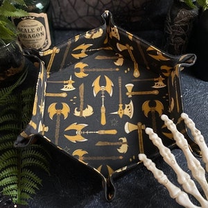 May include: Black hexagonal dice tray with a gold pattern of axes and swords. The tray is made of felt and has a soft, padded interior.