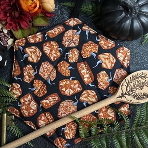 May include: A black and orange fabric pot holder with a pumpkin pattern. The pot holder is lying on a dark surface with a wooden spoon and green ferns.